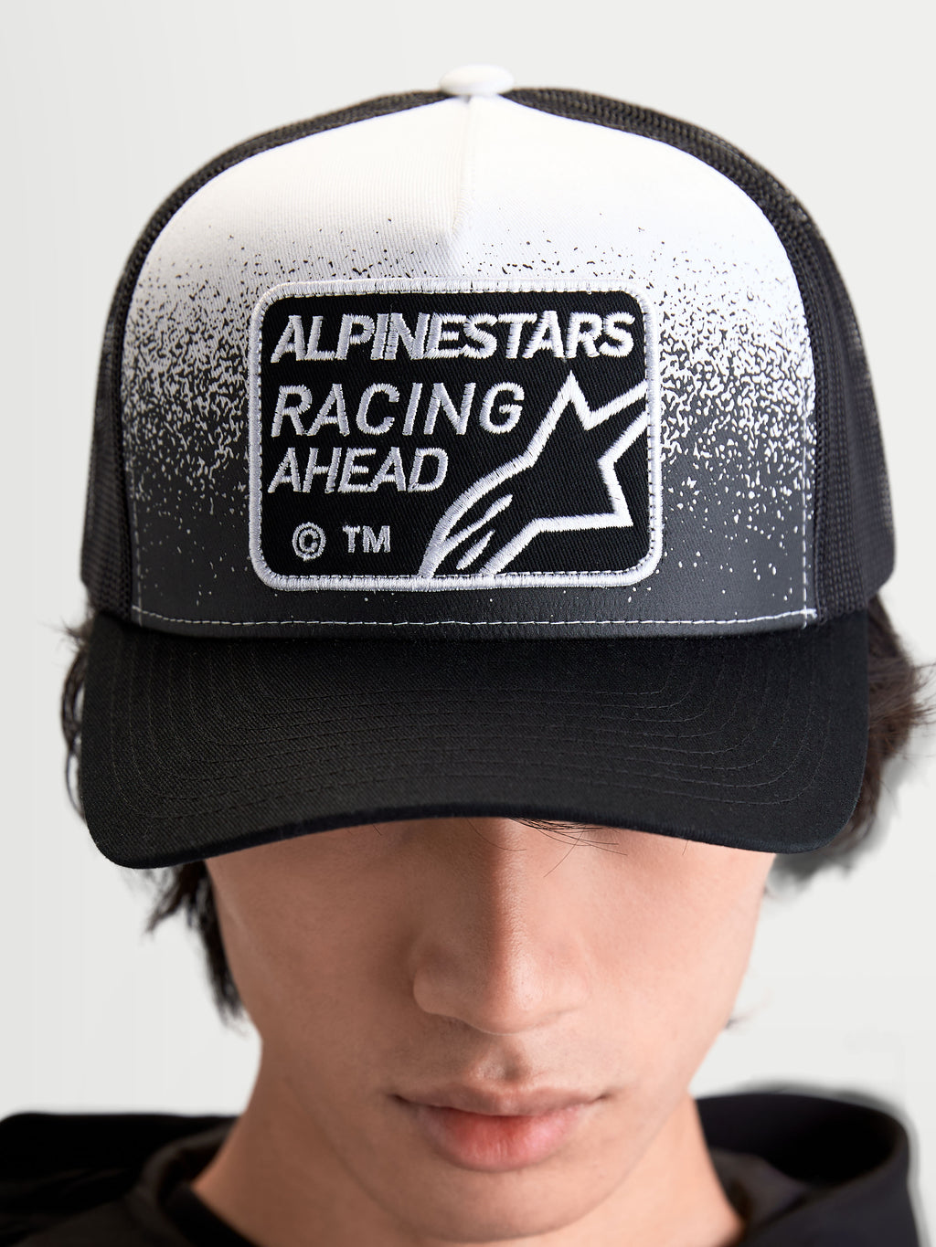 Alpinestars Gravel Trucker pet, sportswear trucker pet voor heren en dames, zwart en wit, met een geweven Alpinestars Racing Ahead-patch met grindtextuurdetails, gestructureerde kroon en gebogen klep, vooraanzicht op model.