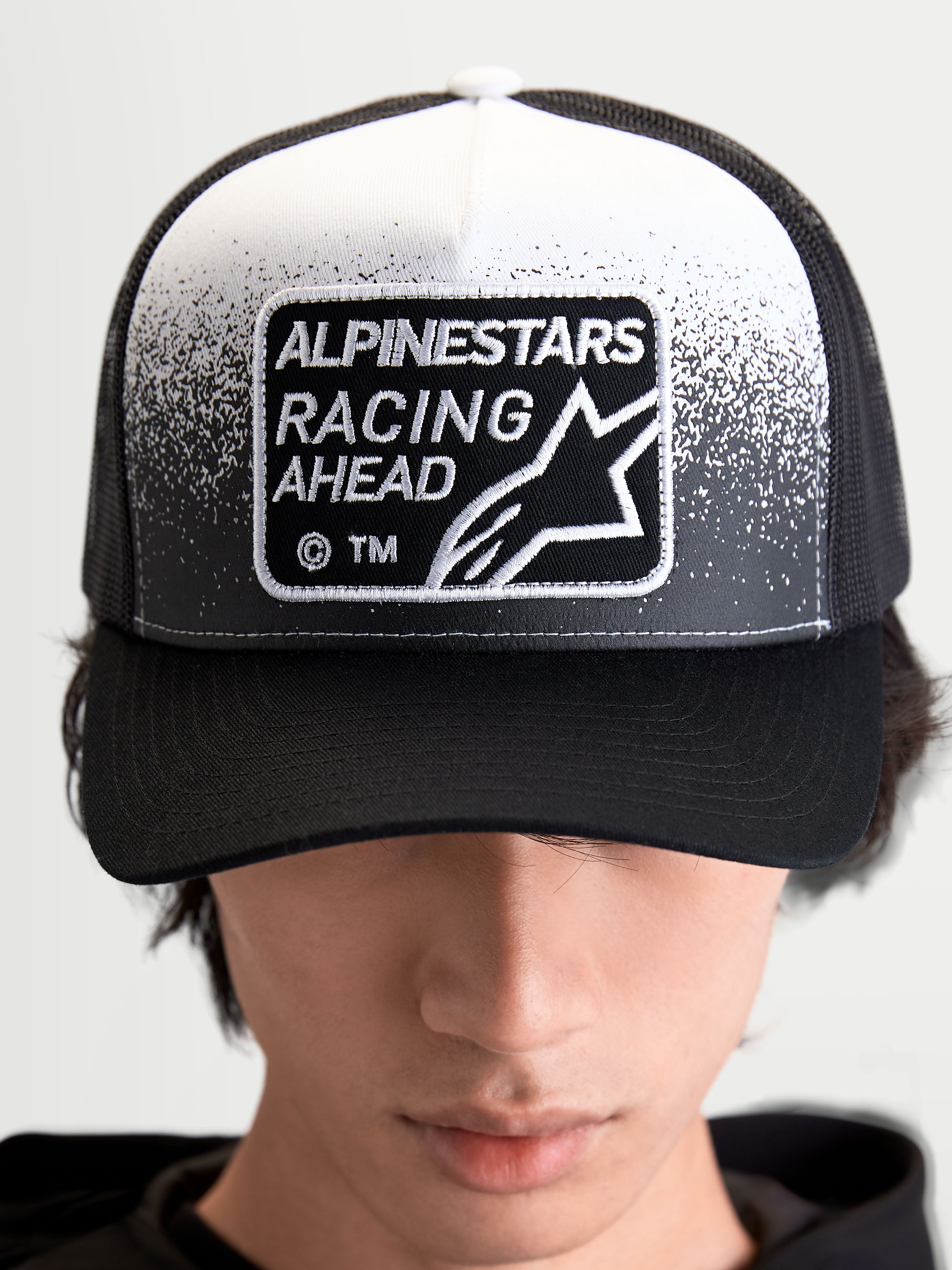 Alpinestars Gravel Trucker pet, sportswear trucker pet voor heren en dames, zwart en wit, met een geweven Alpinestars Racing Ahead-patch met grindtextuurdetails, gestructureerde kroon en gebogen klep, vooraanzicht op model.