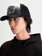 Alpinestars Gravel Trucker pet, casual sportswear pet op model, zwart en wit, voorzien van een gestructureerde kroon, gebogen klep, ademende mesh achterzijde en geweven patch met grindtextuur en Alpinestars Racing Ahead-branding.