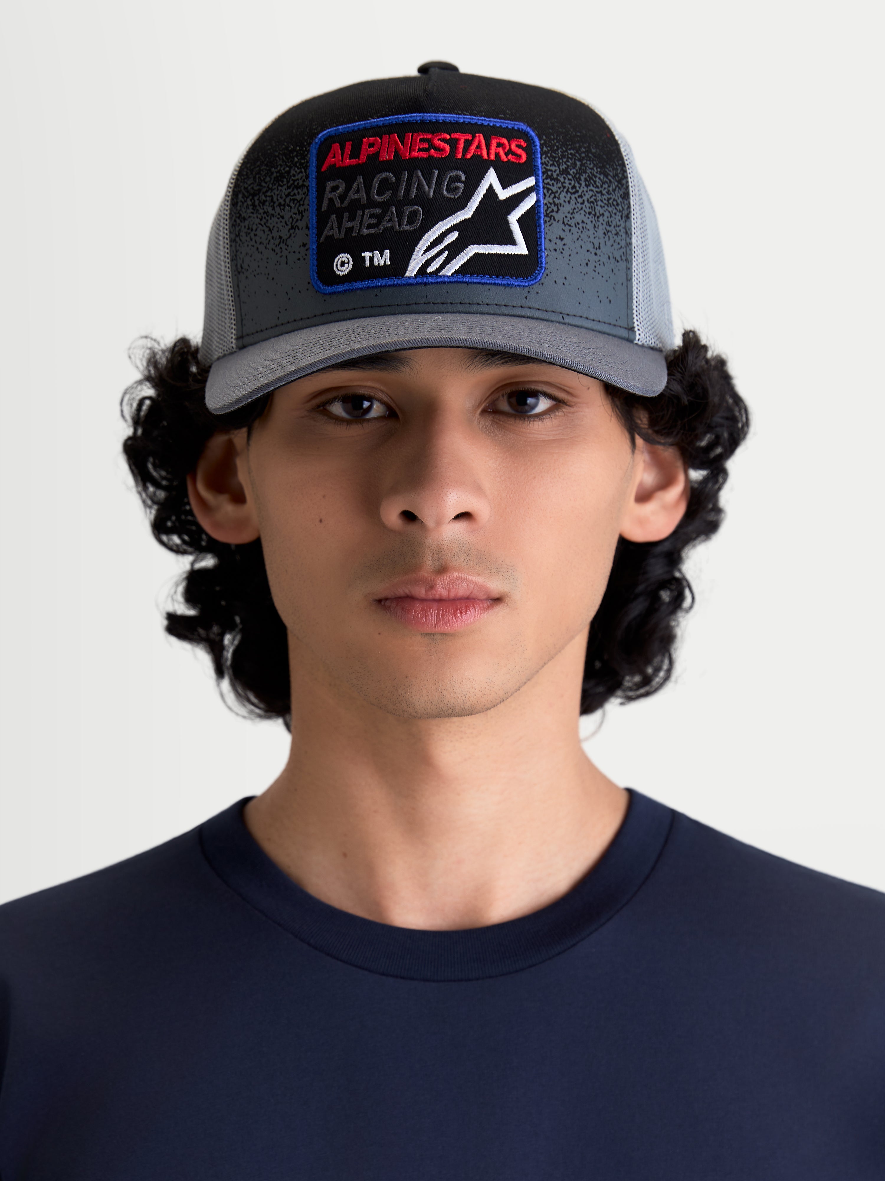 Alpinestars Gravel Trucker pet, casual sportswear met mesh-achterkant voor heren en dames, donkergrijs en zwart, getoond op een model. Voorzien van een gestructureerde kroon, gebogen klep en geweven frontpatch met Alpinestars Racing Ahead-branding en grindtextuurdetails.