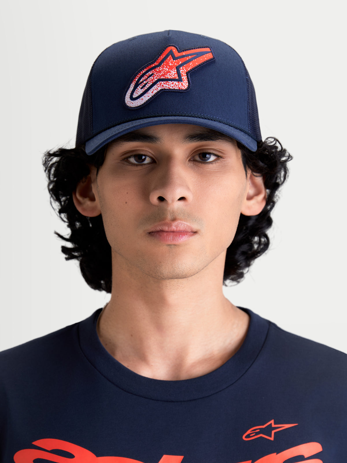 Alpinestars Meteorite Trucker pet, sportswear truckerpet voor heren en dames, marineblauw met rood en wit Astars-logo met grindtextuur, gestructureerde kroon, gebogen klep, vooraanzicht op model