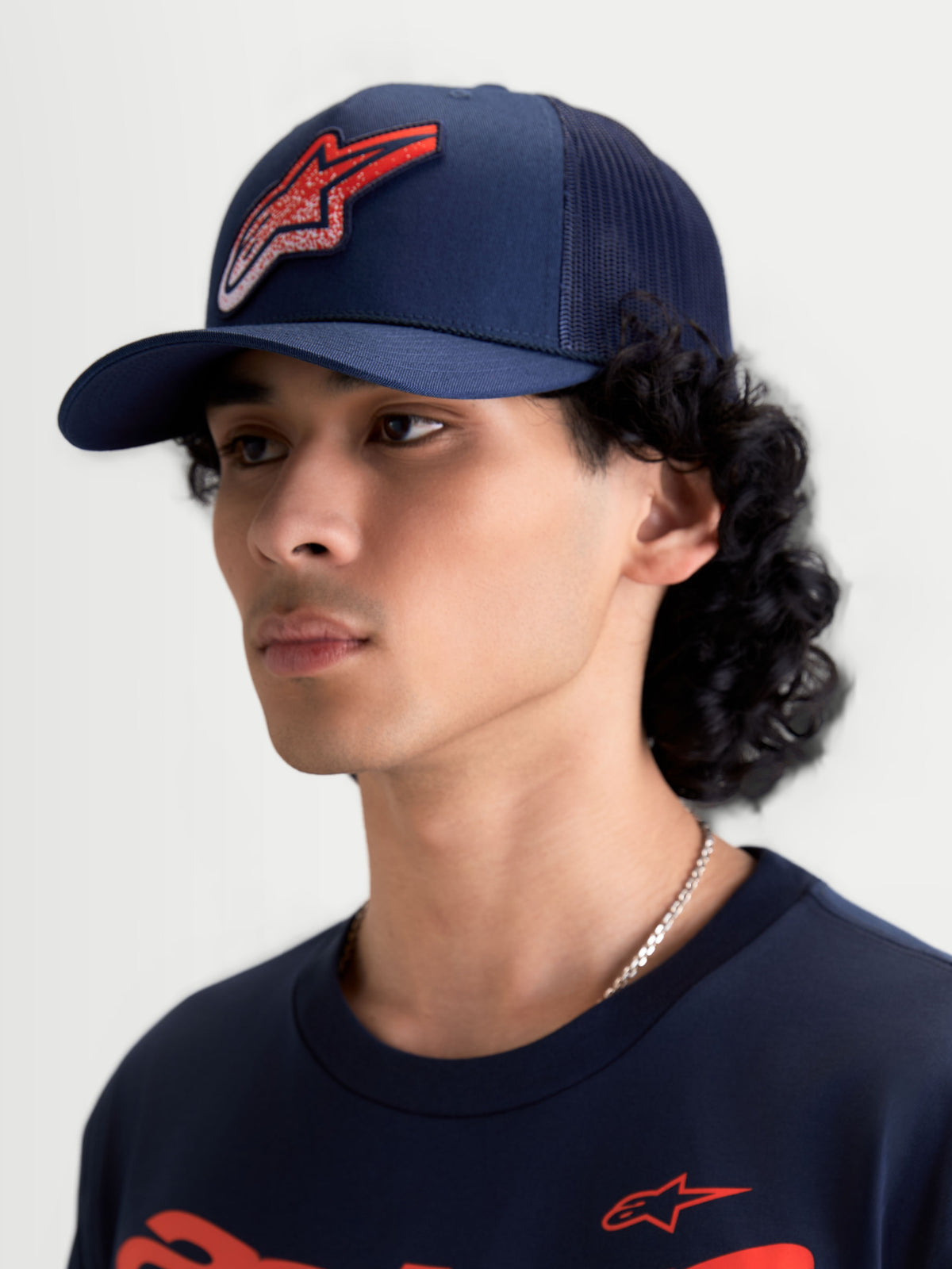 Alpinestars Meteorite Trucker pet, sportswear mesh pet, blauw met rood Astars-logo met grindtextuur, gebogen klep en gestructureerde kroon, gedragen door een model.