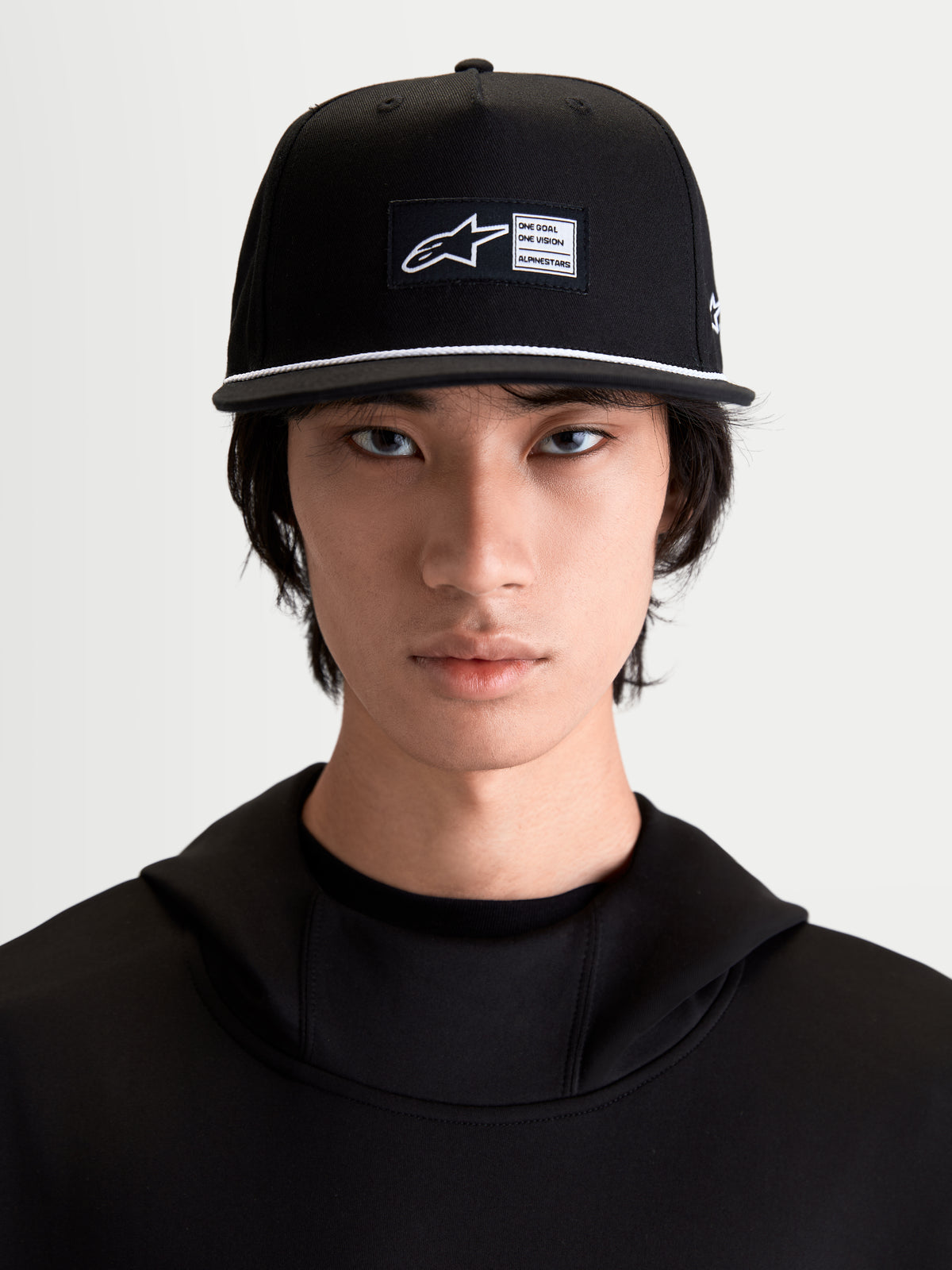 Alpinestars Label Snapback pet, zwarte en witte sportswearpet met platte klep en wit koorddetail, geweven label op de voorkant, katoenen keperstof, op model