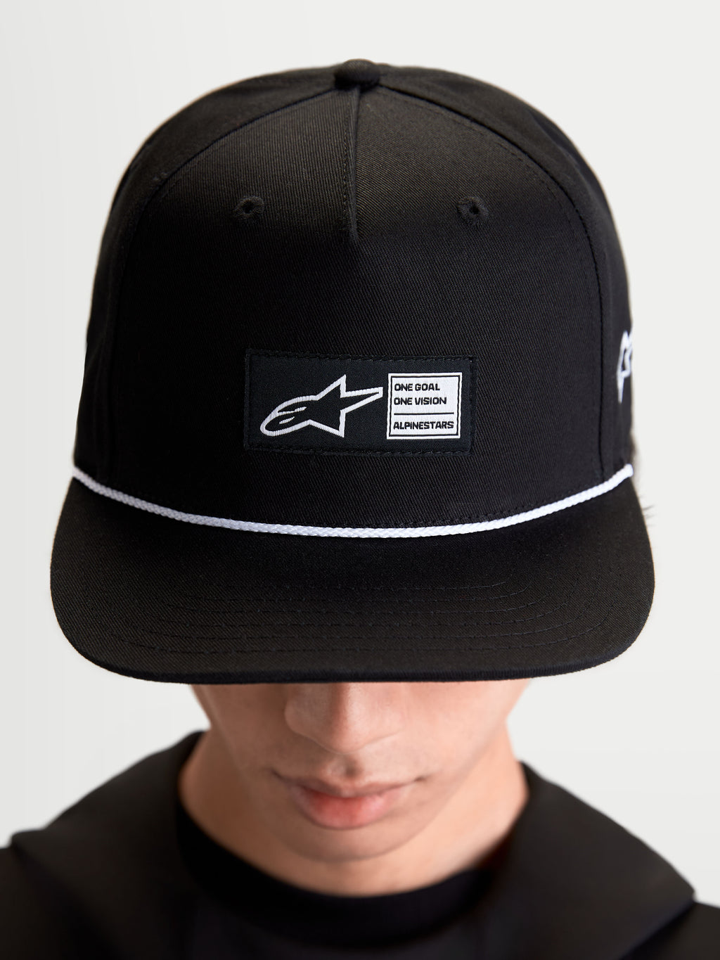 Alpinestars Label Snapback pet, sportswear lifestyle pet, zwart en wit, katoenen keperstof, met een gestructureerde kroon, platte klep, wit koordaccent en een geweven labelpatch aan de voorzijde met Alpinestars-branding, gedragen door een model.