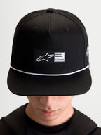 Alpinestars Label Snapback pet, sportswear lifestyle pet, zwart en wit, katoenen keperstof, met een gestructureerde kroon, platte klep, wit koordaccent en een geweven labelpatch aan de voorzijde met Alpinestars-branding, gedragen door een model.