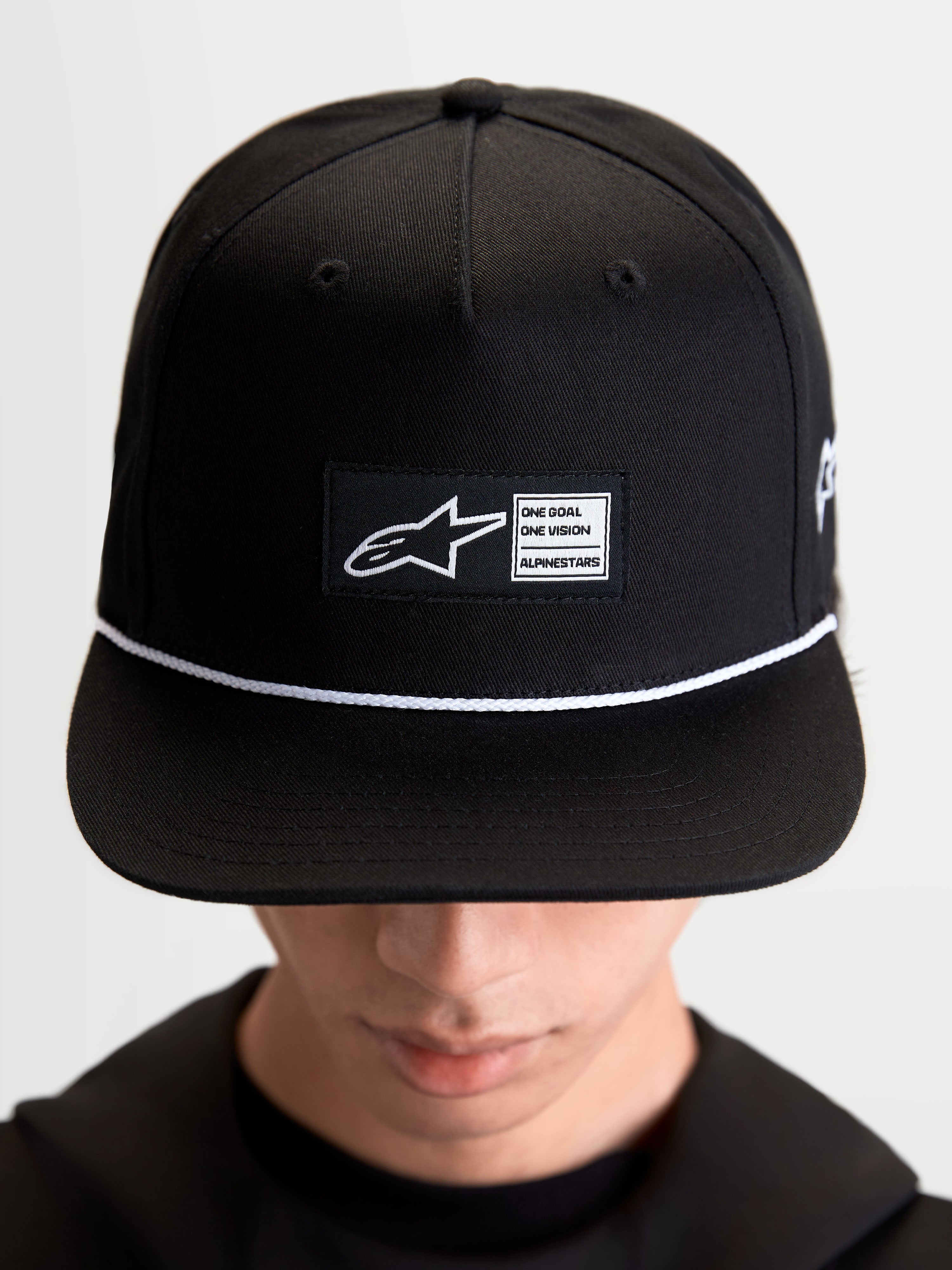 Alpinestars Label Snapback pet, sportswear lifestyle pet, zwart en wit, katoenen keperstof, met een gestructureerde kroon, platte klep, wit koordaccent en een geweven labelpatch aan de voorzijde met Alpinestars-branding, gedragen door een model.