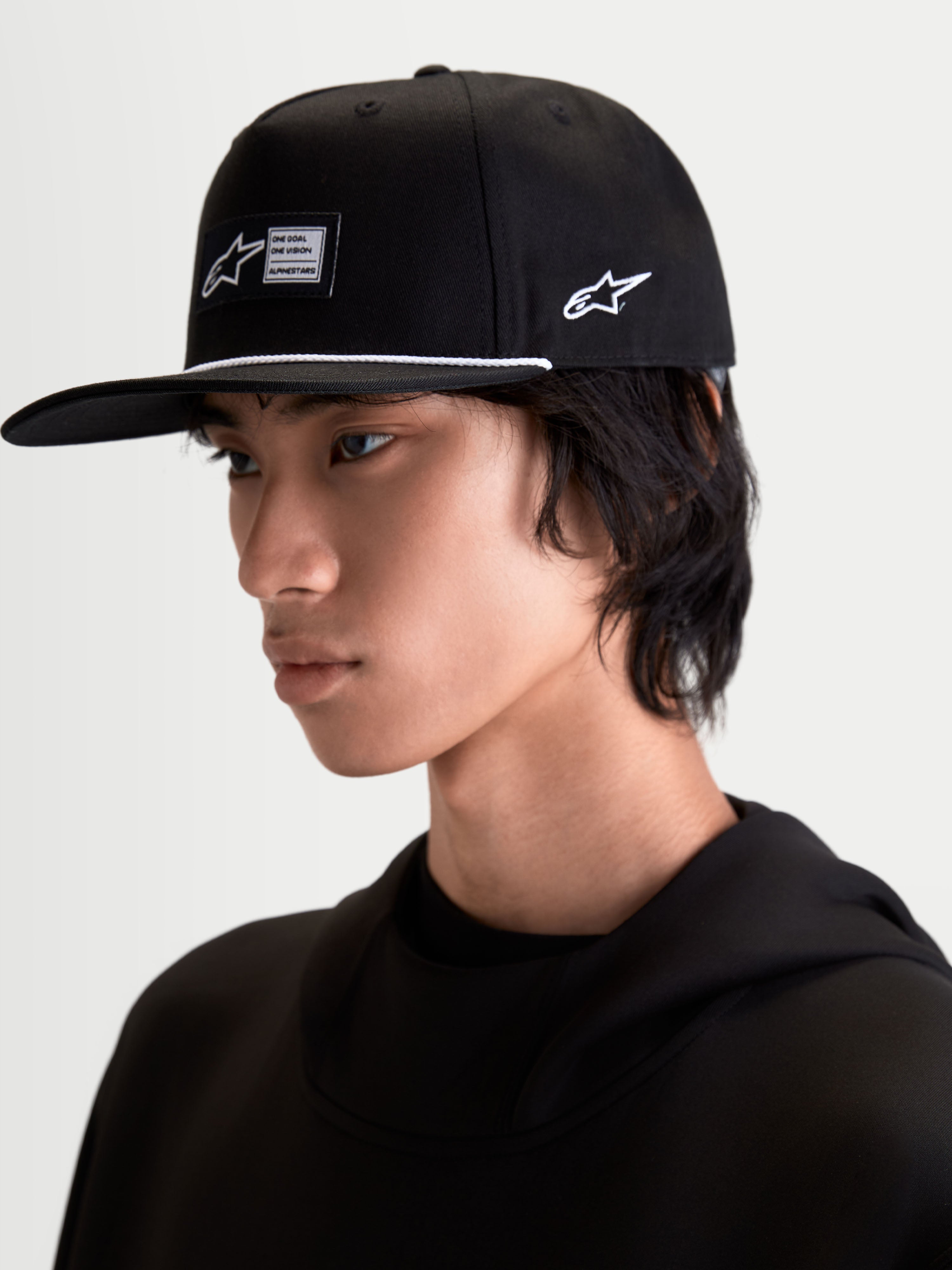 Alpinestars Label Snapback pet, zwarte sportswearpet met platte klep en gestructureerde kroon, voorzien van een geweven label aan de voorzijde, wit accentkoord en zijlogo-borduurwerk, getoond op een model voor een moderne motorsport-geïnspireerde lifestyle-look.