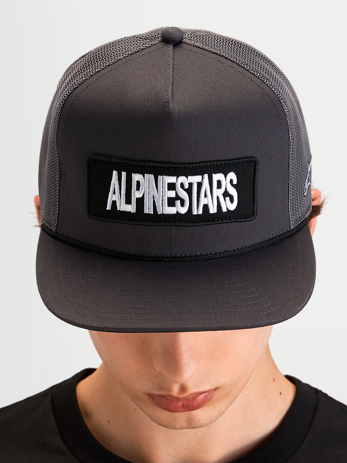 Alpinestars Ten Trucker pet, sportieve pet met platte klep gedragen door model, donkergrijs, met Alpinestars woordlogoborduurpatch op de voorkant en gestructureerde mesh trucker kroon