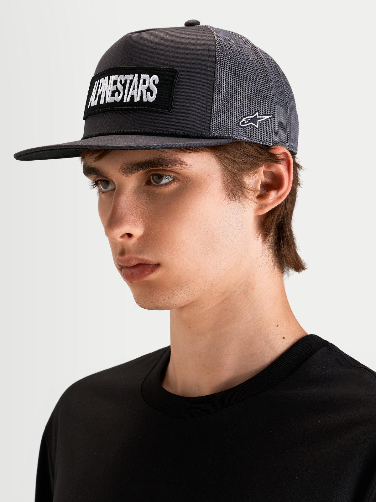 Alpinestars Ten Trucker pet, lifestyle pet met platte klep in donkergrijs, gedragen door model, met geborduurd Alpinestars woordlogo op de voorkant en Astars-logo op de zijkant