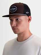Alpinestars Hotlink Trucker pet op model, bruine en zwarte lifestyle sportswearpet met platte klep, gestructureerde kroon van twill en een geweven Astars-logopatch met fietskettingrand.