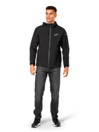Alpinestars Kascade regenjas, heren regenjas, zwart, gedragen met donkergrijze jeans en zwarte sneakers, voorzien van een ontwerp met capuchon, gecoate rits aan de voorzijde en wit Alpinestars-logo op de borst, casual motorsport-geïnspireerde streetwear