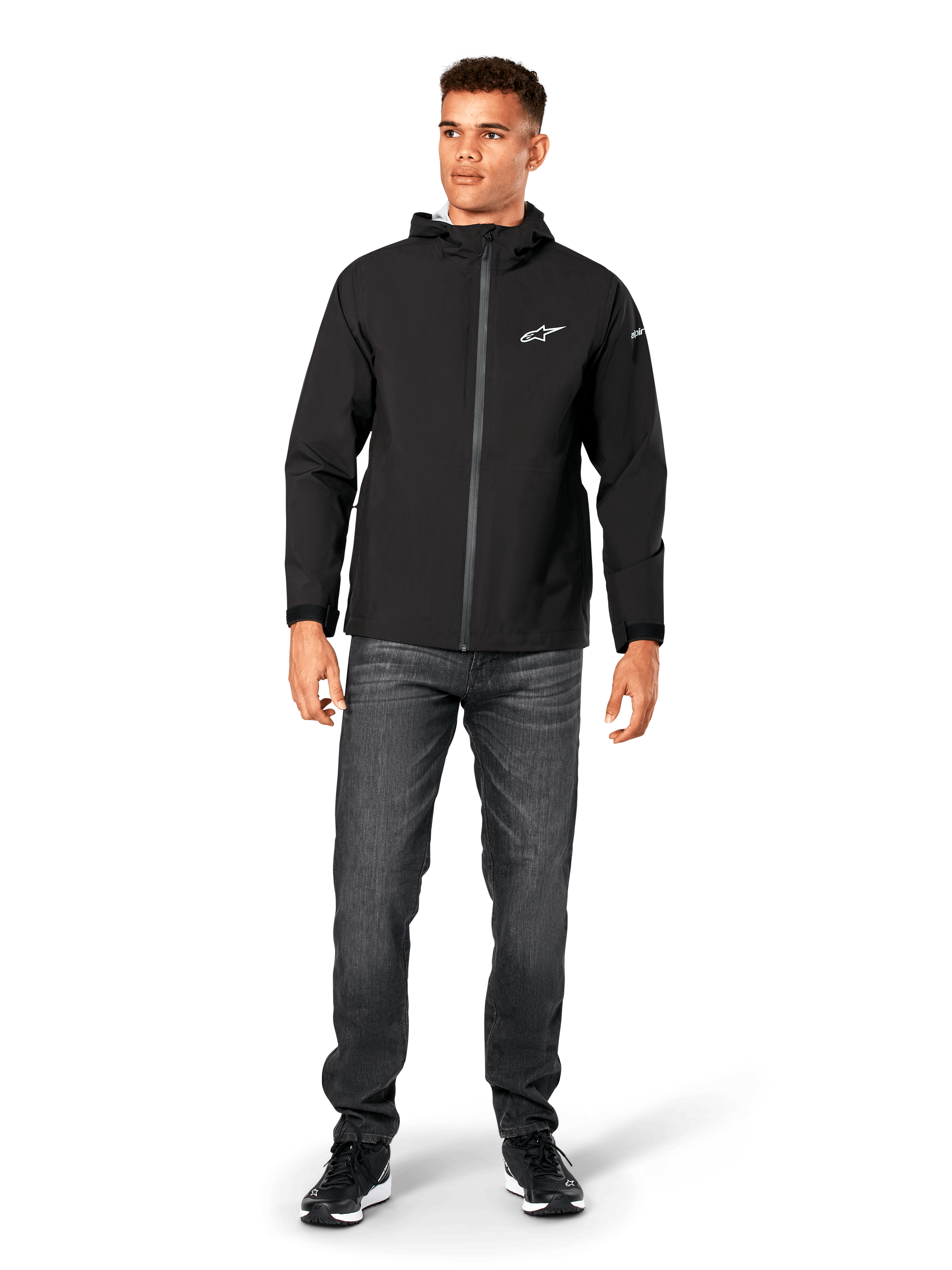 Alpinestars Kascade regenjas, heren regenjas, zwart, gedragen met donkergrijze jeans en zwarte sneakers, voorzien van een ontwerp met capuchon, gecoate rits aan de voorzijde en wit Alpinestars-logo op de borst, casual motorsport-geïnspireerde streetwear