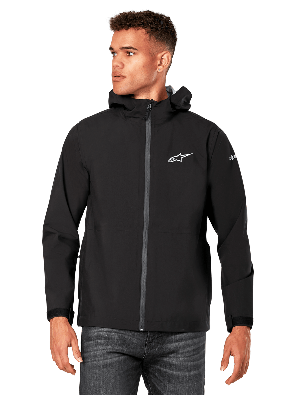 Mannelijk model draagt Alpinestars Kascade regenjas, zwart, wit logo op de borst en merknaam op de mouw, regenjas met capuchon met gecoate rits aan de voorzijde en klittenbandmanchetten, gecombineerd met een donkere jeans, casual motorsport-geïnspireerde streetwear