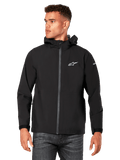 Mannelijk model draagt Alpinestars Kascade regenjas, zwart, wit logo op de borst en merknaam op de mouw, regenjas met capuchon met gecoate rits aan de voorzijde en klittenbandmanchetten, gecombineerd met een donkere jeans, casual motorsport-geïnspireerde streetwear