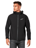 Mannelijk model draagt Alpinestars Kascade regenjas, zwart, wit logo op de borst en merknaam op de mouw, regenjas met capuchon met gecoate rits aan de voorzijde en klittenbandmanchetten, gecombineerd met een donkere jeans, casual motorsport-geïnspireerde streetwear