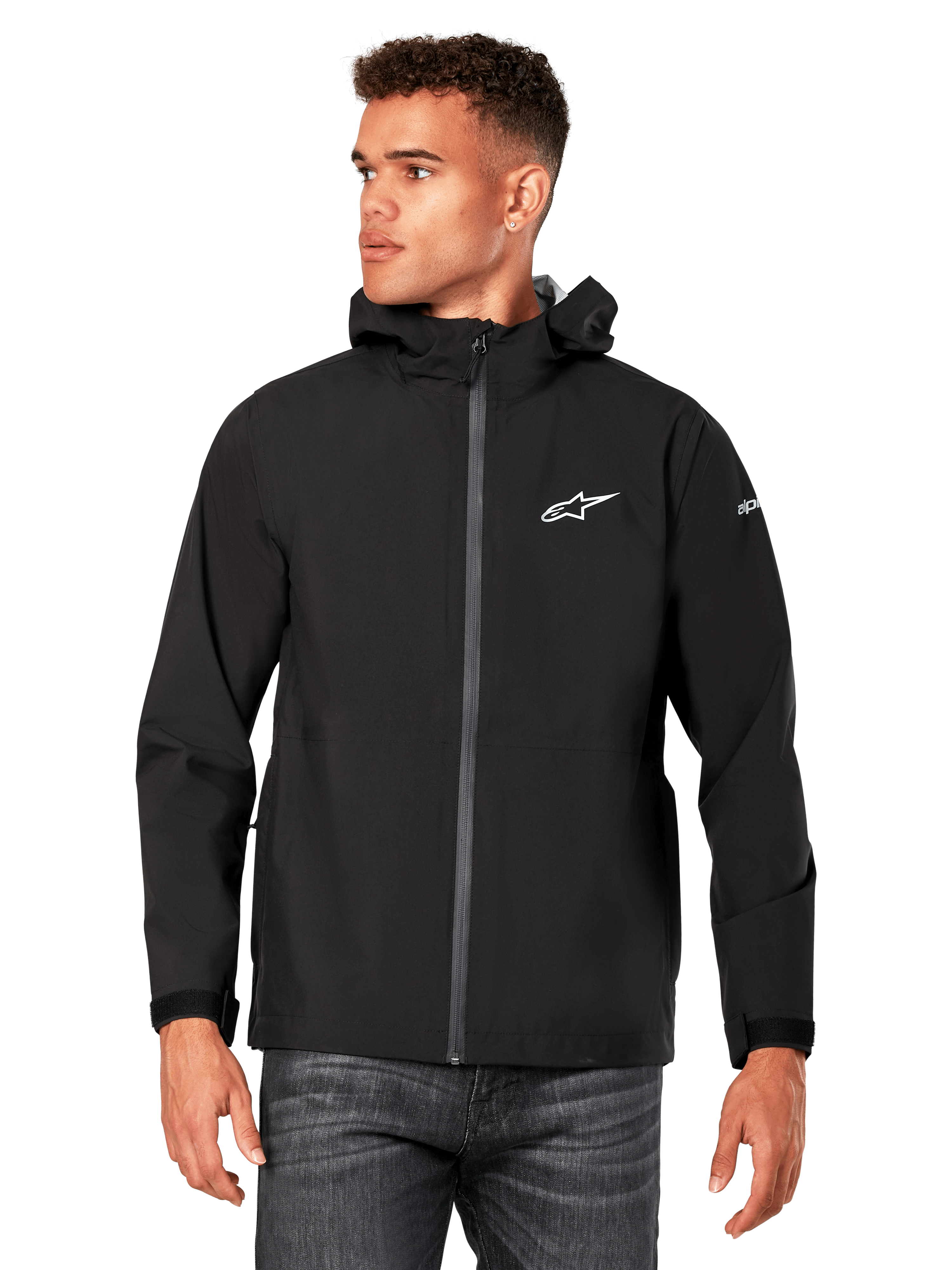 Mannelijk model draagt Alpinestars Kascade regenjas, zwart, wit logo op de borst en merknaam op de mouw, regenjas met capuchon met gecoate rits aan de voorzijde en klittenbandmanchetten, gecombineerd met een donkere jeans, casual motorsport-geïnspireerde streetwear