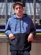 Alpinestars Speeder Windbreaker jas, leigrijs blauw en zwart, windbreaker met capuchon en kleurblok-ontwerp, wit logo op de borst, casual lifestylekleding gedragen op een circuit