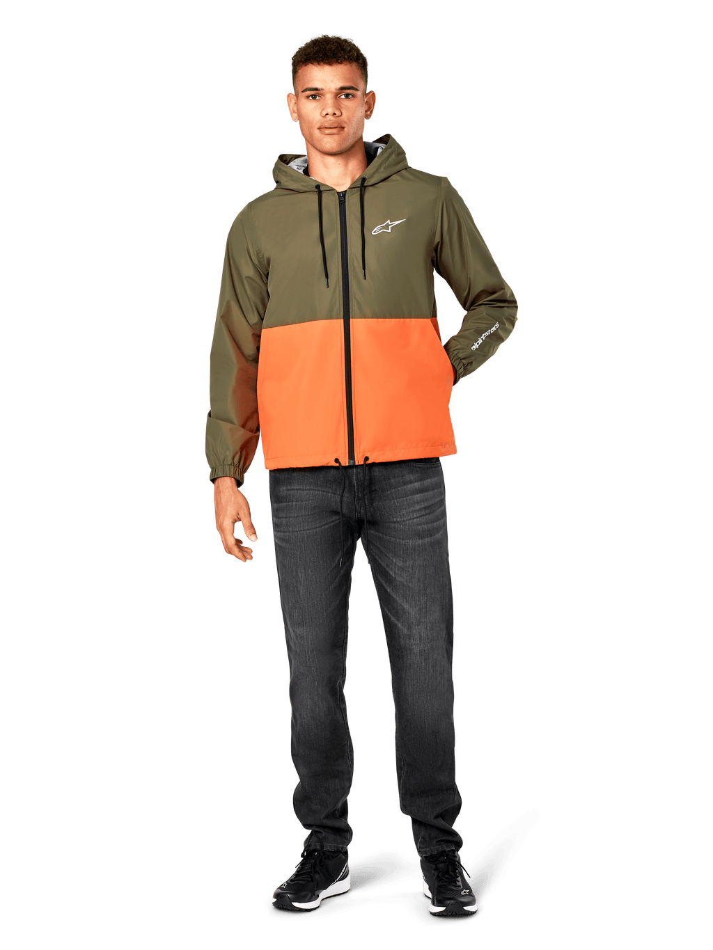 Alpinestars Speeder Windbreaker jas, kleurblokken in Military Green en oranje, lichtgewicht jas met capuchon, zwarte trekkoorden en ritssluiting, wit logo op de borst en mouw, gedragen met donkere jeans en zwarte sneakers, casual sportswear stijl.