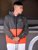 Alpinestars Speeder Windbreaker Jas, military orange en donkergroen color-block design, vrijetijdsjas met capuchon, witte logo's op de borst en mouw, gecombineerd met een witte broek voor een op de motorsport geïnspireerde lifestyle-look