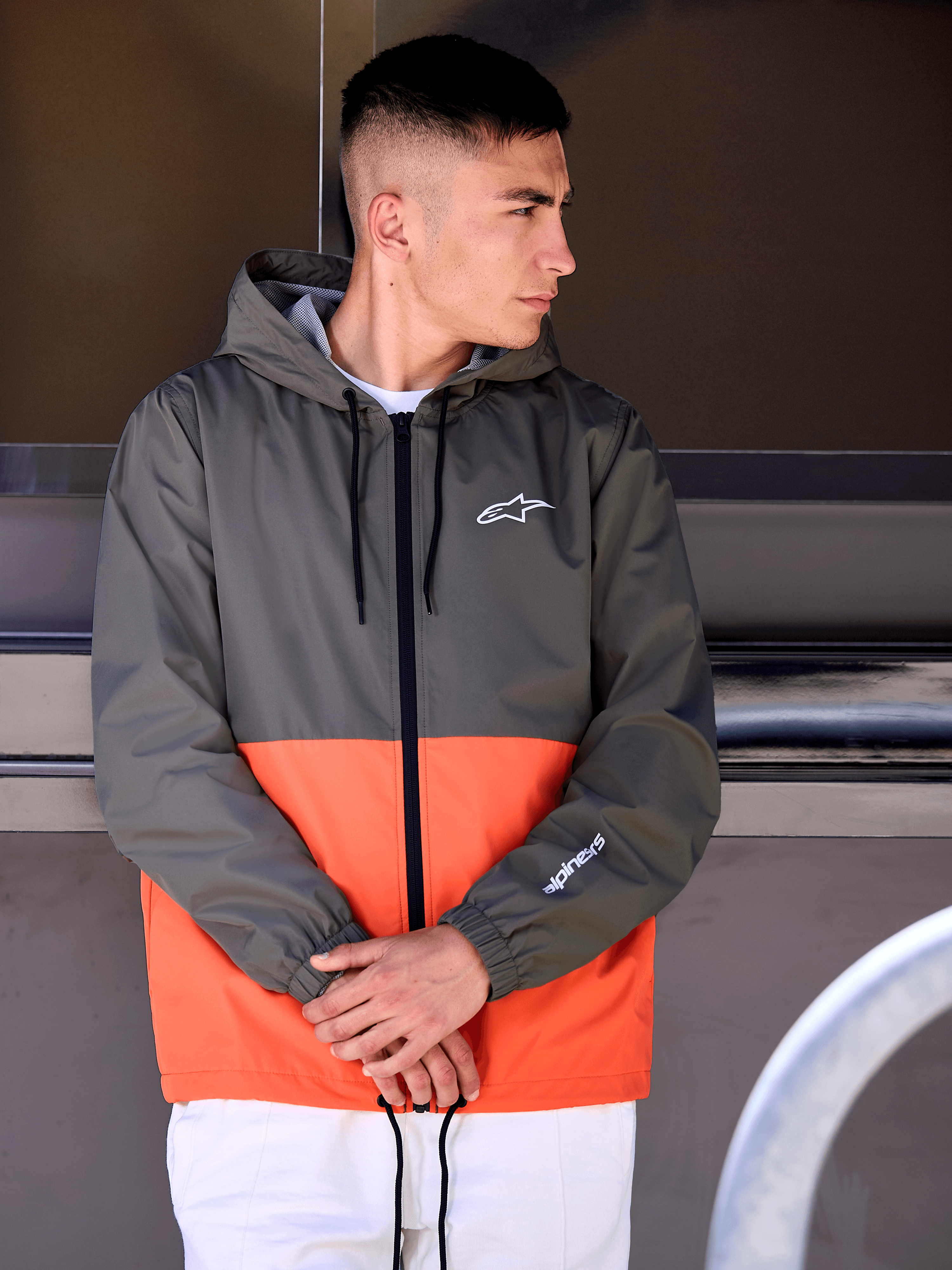 Alpinestars Speeder Windbreaker Jas, military orange en donkergroen color-block design, vrijetijdsjas met capuchon, witte logo's op de borst en mouw, gecombineerd met een witte broek voor een op de motorsport geïnspireerde lifestyle-look