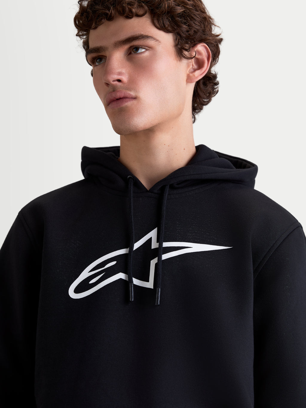 Alpinestars Ageless V3 Hoodie, heren-sportswearpullover van fleece, zwart en wit, close-up op model met een groot wit Alpinestars-logo op de borst en een capuchon met trekkoord.