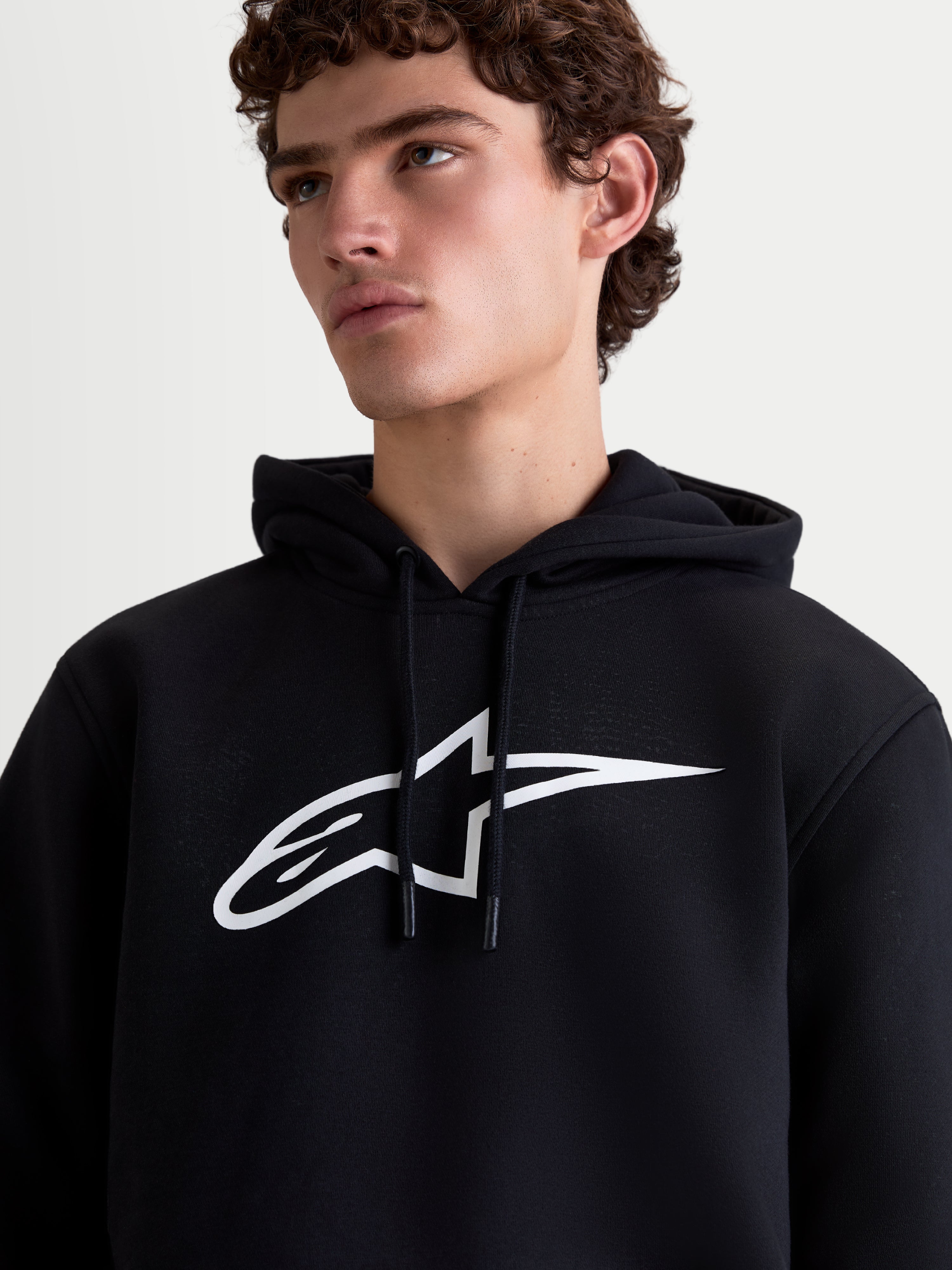 Alpinestars Ageless V3 Hoodie, heren-sportswearpullover van fleece, zwart en wit, close-up op model met een groot wit Alpinestars-logo op de borst en een capuchon met trekkoord.