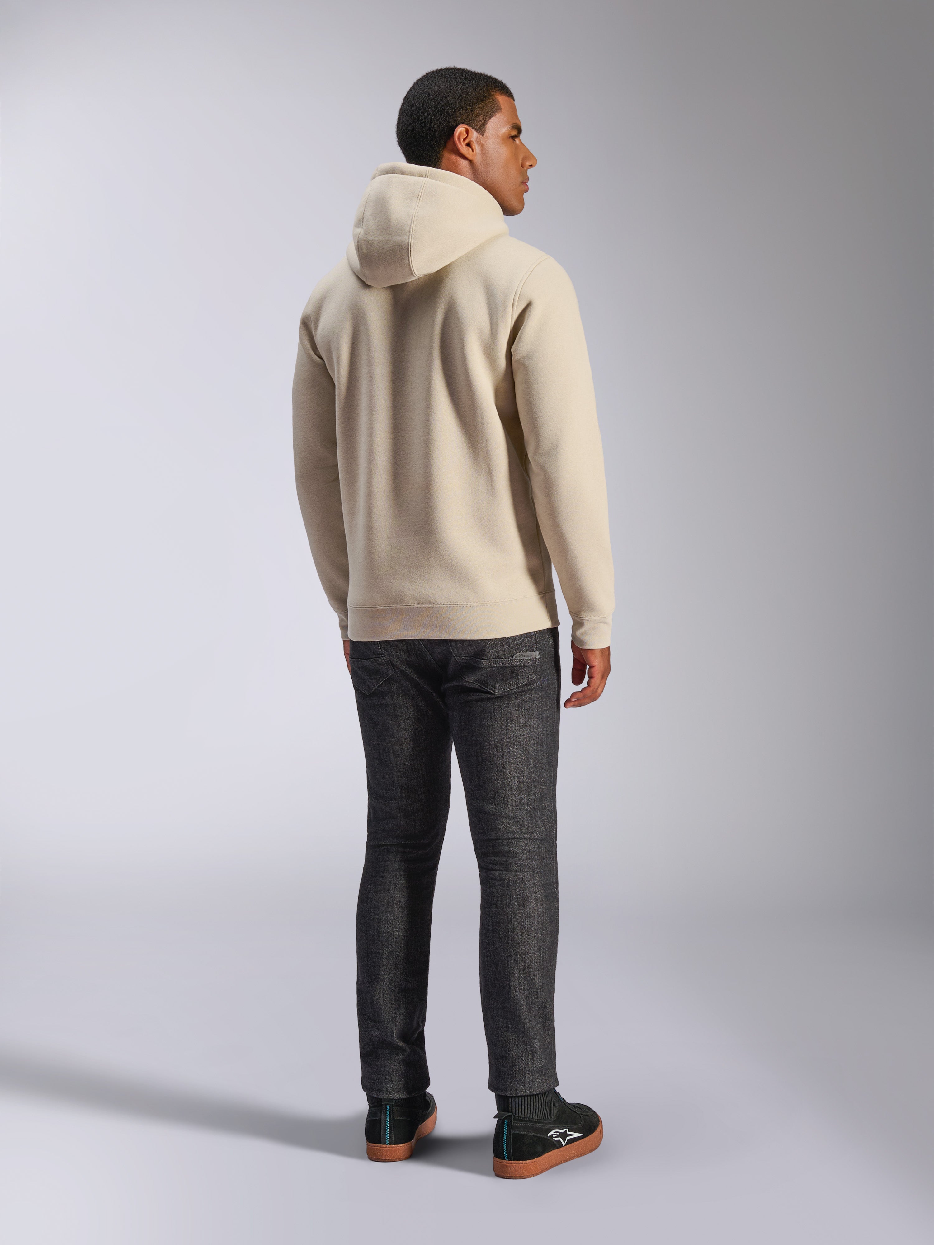 Alpinestars Ageless V3 Hoodie, casual off-white fleece trui met capuchon van de achterkant gezien, voorzien van geribde manchetten en zoom, gedragen met een donkere slim-fit jeans en zwarte Alpinestars sneakers
