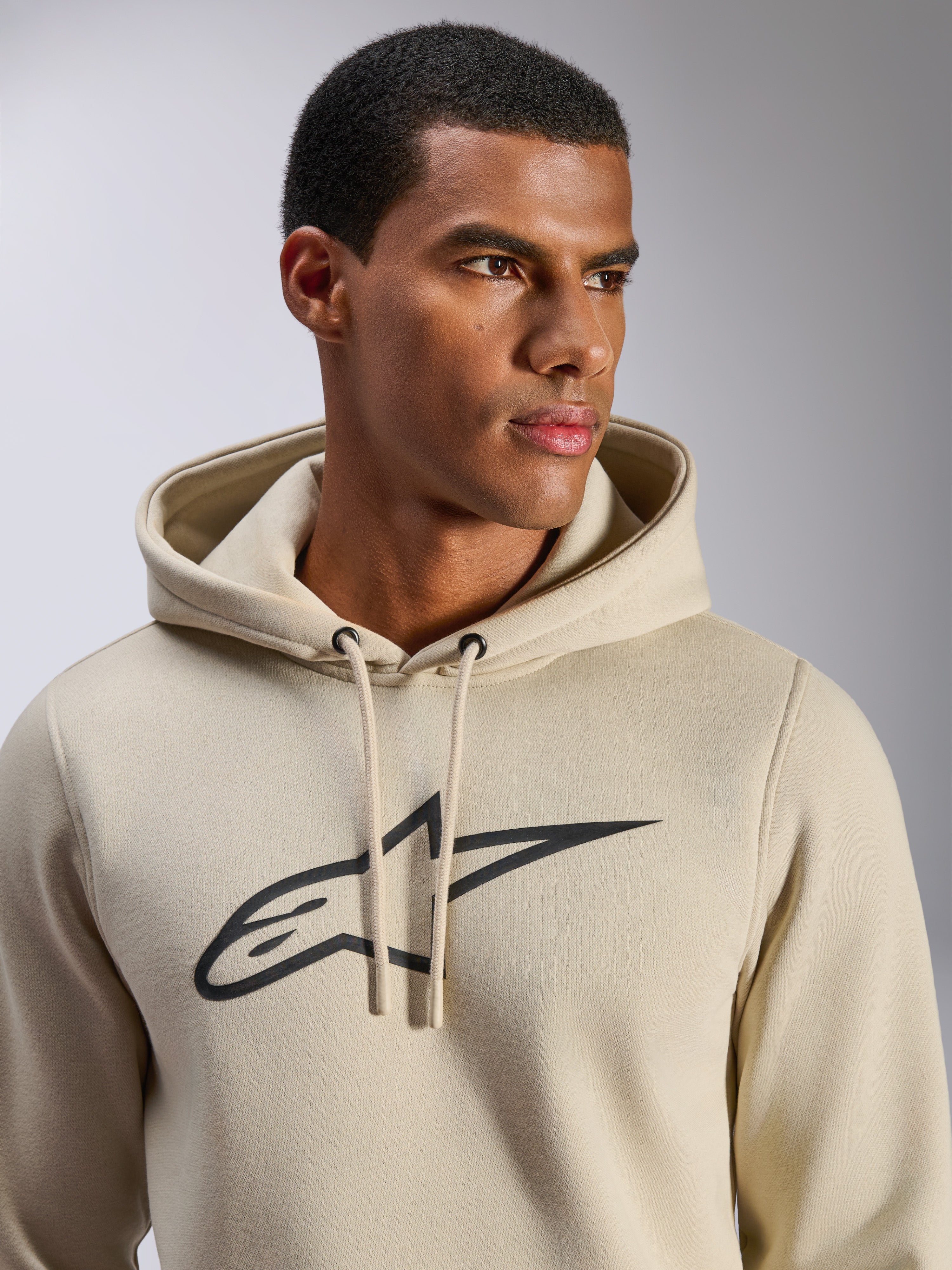 Alpinestars Ageless V3 hoodie, casual sportkleding, gebroken wit met zwart geborduurd Alpinestars-sterlogo, capuchon met trekkoord, close-up op model, zachte fleece, modern op de motorsport geïnspireerd ontwerp