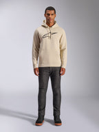 Alpinestars Ageless V3 Hoodie, casual sportkleding, Off White met zwart geborduurd logo, gedragen door een man met een donkergrijze broek en zwarte sneakers, voorzien van een capuchon met trekkoord en kangoeroezak, modern op de motorsport geïnspireerd ontwerp