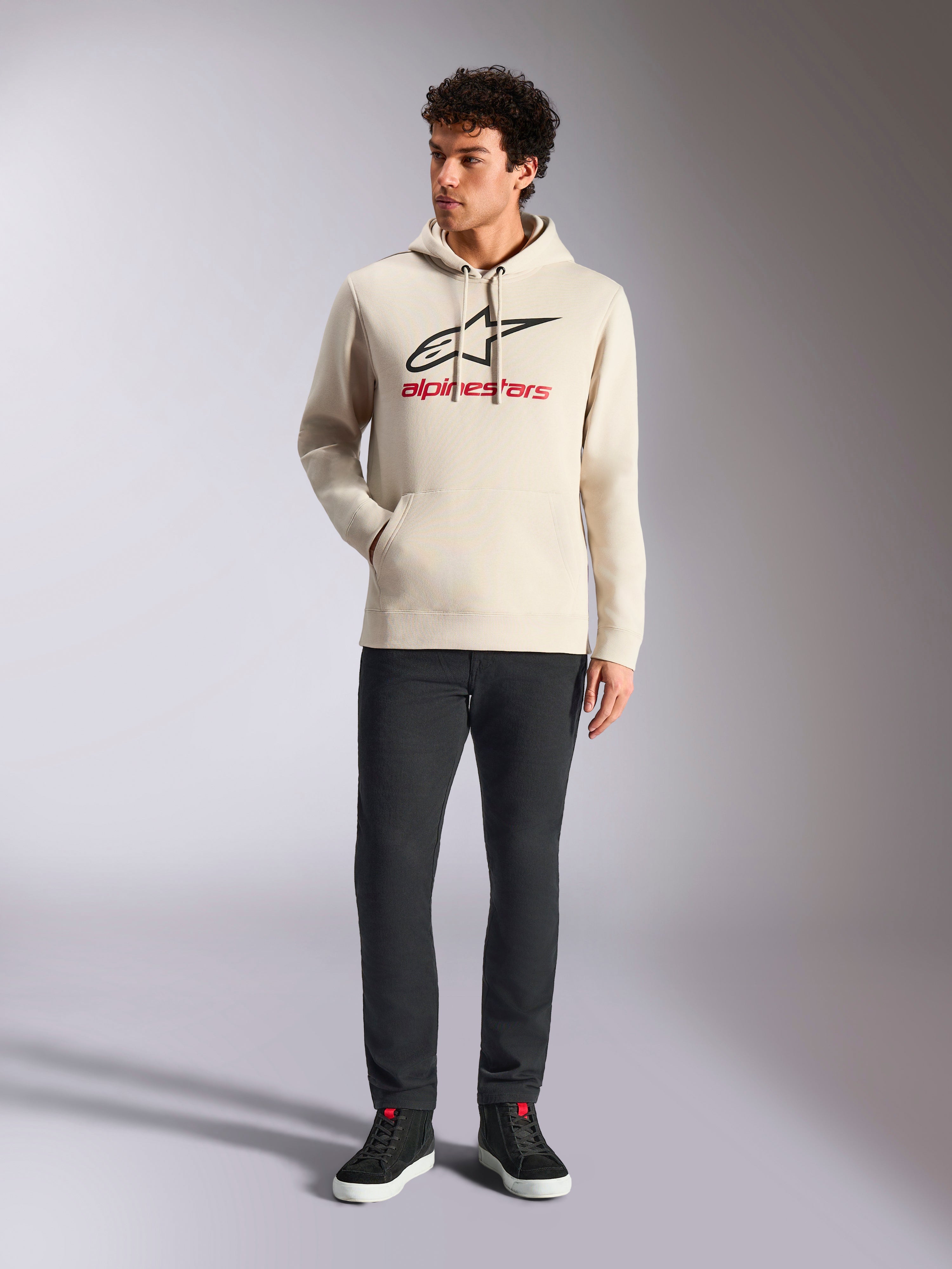 Alpinestars Always V3 Hoodie, casual streetwear, gebroken wit met zwart sterlogo en rode belettering, capuchon met trekkoord, lange mouwen, gedragen door een mannelijk model met een zwarte broek en zwarte sneakers
