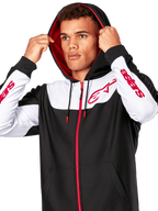 Alpinestars Sessions V3 Hoodie, zwart wit en rood, hoodie met ritssluiting gedragen door een man met de capuchon op, rode Alpinestars-logo's op de borst en mouwen, casual motorsport streetwear