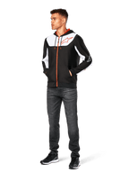 Alpinestars Sessions V3 Hoodie, casual hoodie met ritssluiting, zwart, wit en rood, gedragen door model met donkere jeans en zwarte sneakers, voorzien van witte kleurvlakken op de borst en mouwen, rode rits- en trekkoorddetails, groot logo op de borst, poly-fleece motorsport-geïnspireerde streetwear