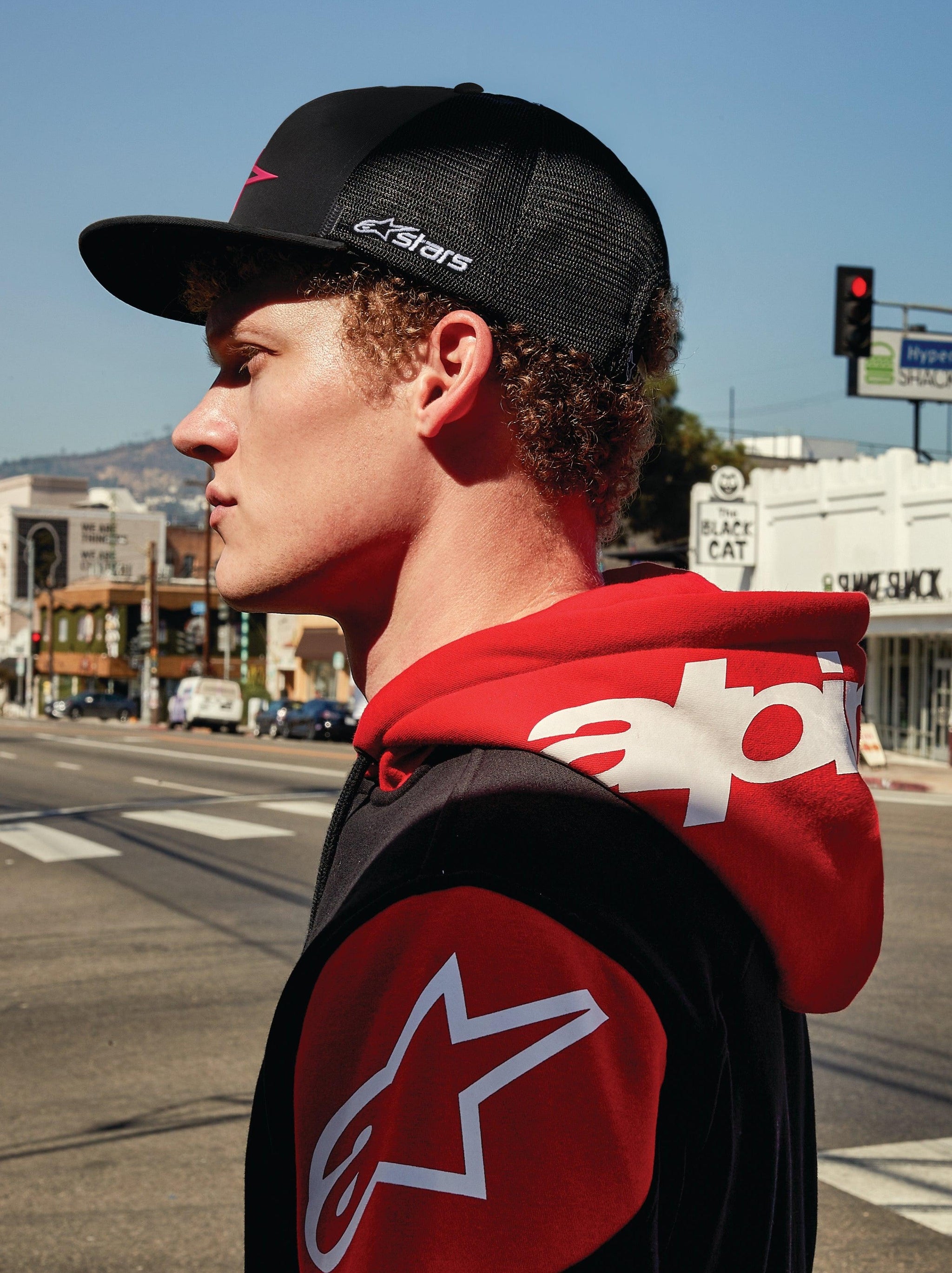 Alpinestars 3D Ageless Trucker Hat, zwarte trucker pet met mesh achterkant en rood logo, gedragen met een rood-zwarte Alpinestars hoodie met opvallende witte logo's, zijprofiel van een man in een stedelijke omgeving, casual motorsport streetwear