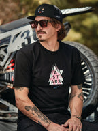 Alpinestars Gambit Trucker pet in zwart met ronde logopatch, gedragen met Alpinestars Soloist SS Tee in zwart, lifestyle-foto van een man met zonnebril en tatoeages zittend voor een motorcrossmotor