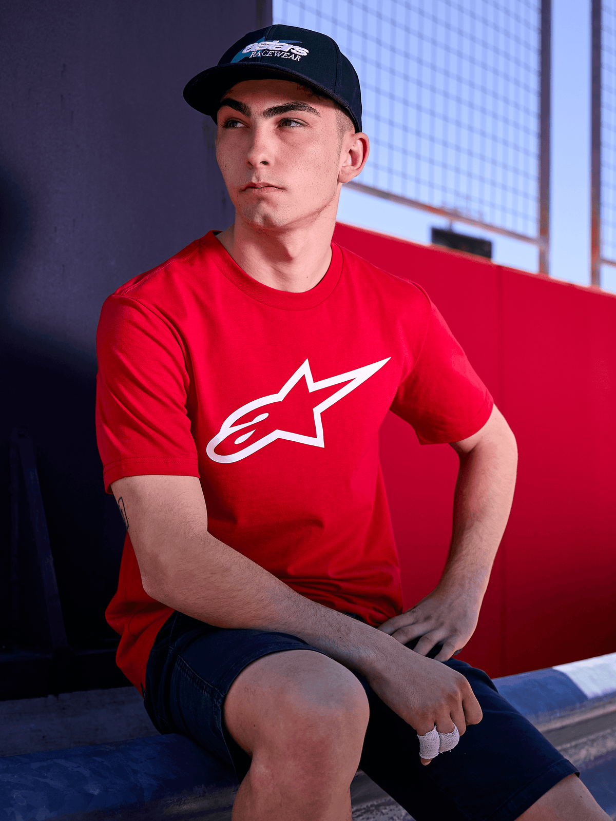 Alpinestars Profile pet in het zwart, lifestyle-opname van een man die de pet met gebogen klep draagt, gecombineerd met een rood t-shirt met een wit Alpinestars-logo, casual op motorsport geïnspireerde streetwear