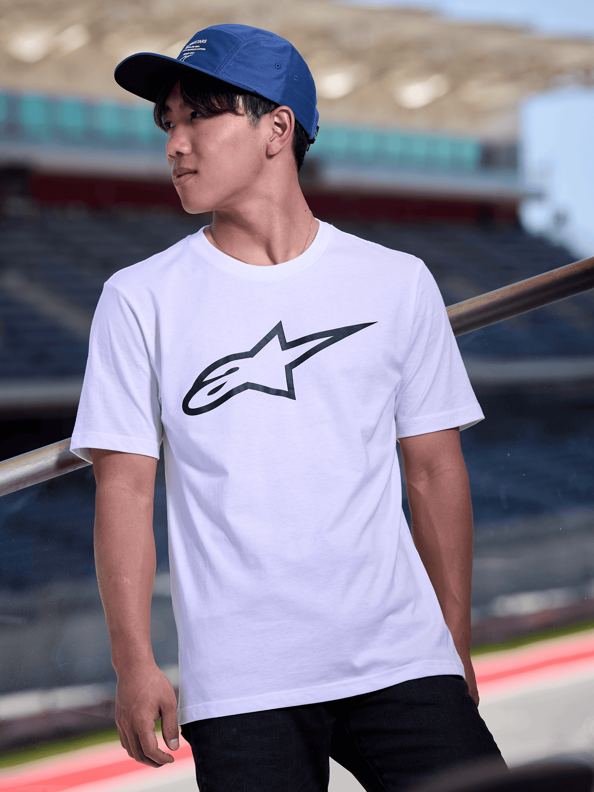 Man met Alpinestars Peasy pet in blauw, 5-panel jockey pet met platte klep en witte grafische logo-opdruk, gecombineerd met een wit Alpinestars logo T-shirt, casual motorsport-geïnspireerde streetwear