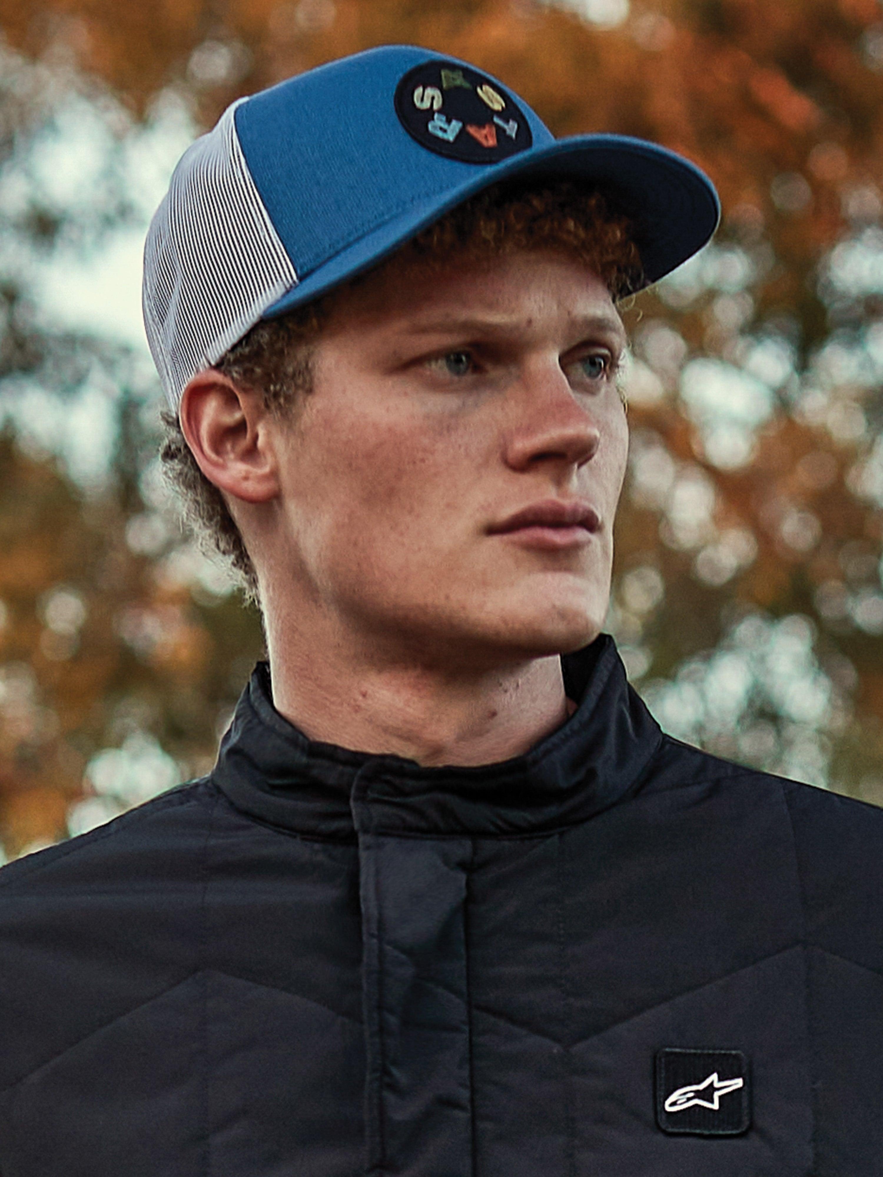 Man met Alpinestars Gambit Trucker pet, blauw-witte cap met mesh-achterkant en een ronde Astars logo patch, gecombineerd met een zwart gewatteerd Alpinestars jack, casual motorsport-ge&iuml;nspireerd lifestyle portret met herfstachtergrond