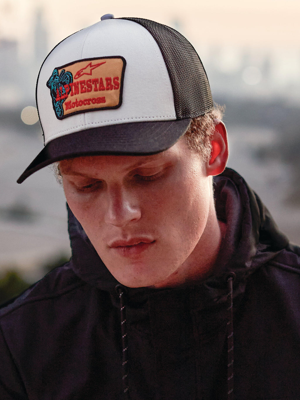Alpinestars Hardware Trucker pet in Military White, gedragen door een man in een zwarte hoodie, voorzien van een wit voorpaneel met een zwarte gebogen klep en zwarte mesh achterkant, beige geborduurde patch met rode Alpinestars Motocross-branding, casual lifestyle motorsport streetwear