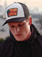 Alpinestars Hardware Trucker pet in Military White, gedragen door een man in een zwarte hoodie, voorzien van een wit voorpaneel met een zwarte gebogen klep en zwarte mesh achterkant, beige geborduurde patch met rode Alpinestars Motocross-branding, casual lifestyle motorsport streetwear