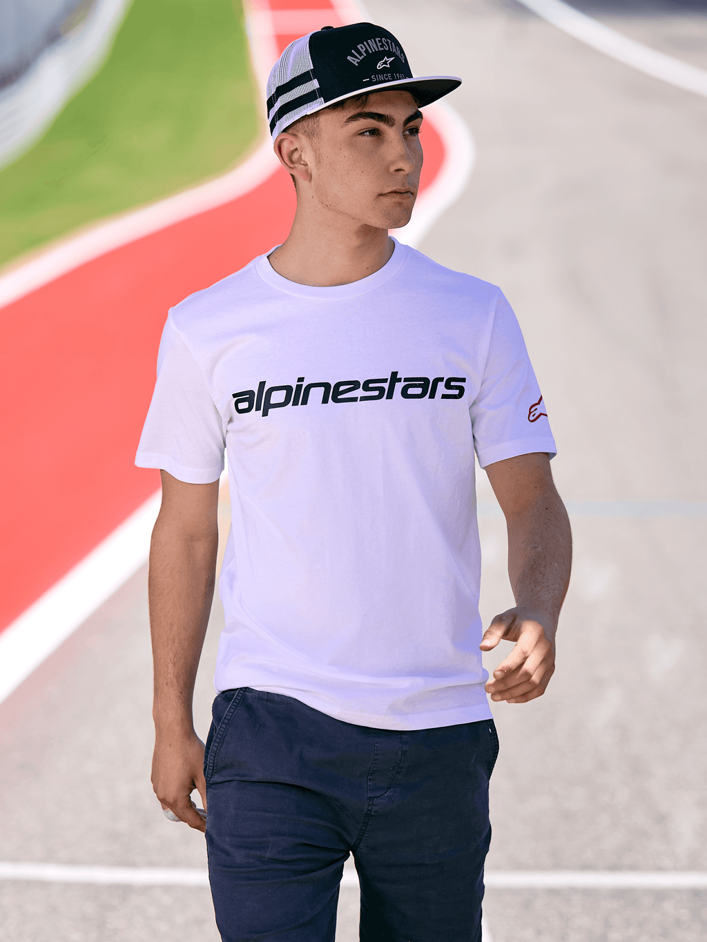 Alpinestars Backline Trucker Pet in zwart en wit, man draagt een wit Alpinestars logo T-shirt en een donkerblauwe broek, casual motorsport streetwear op een buitenbaan