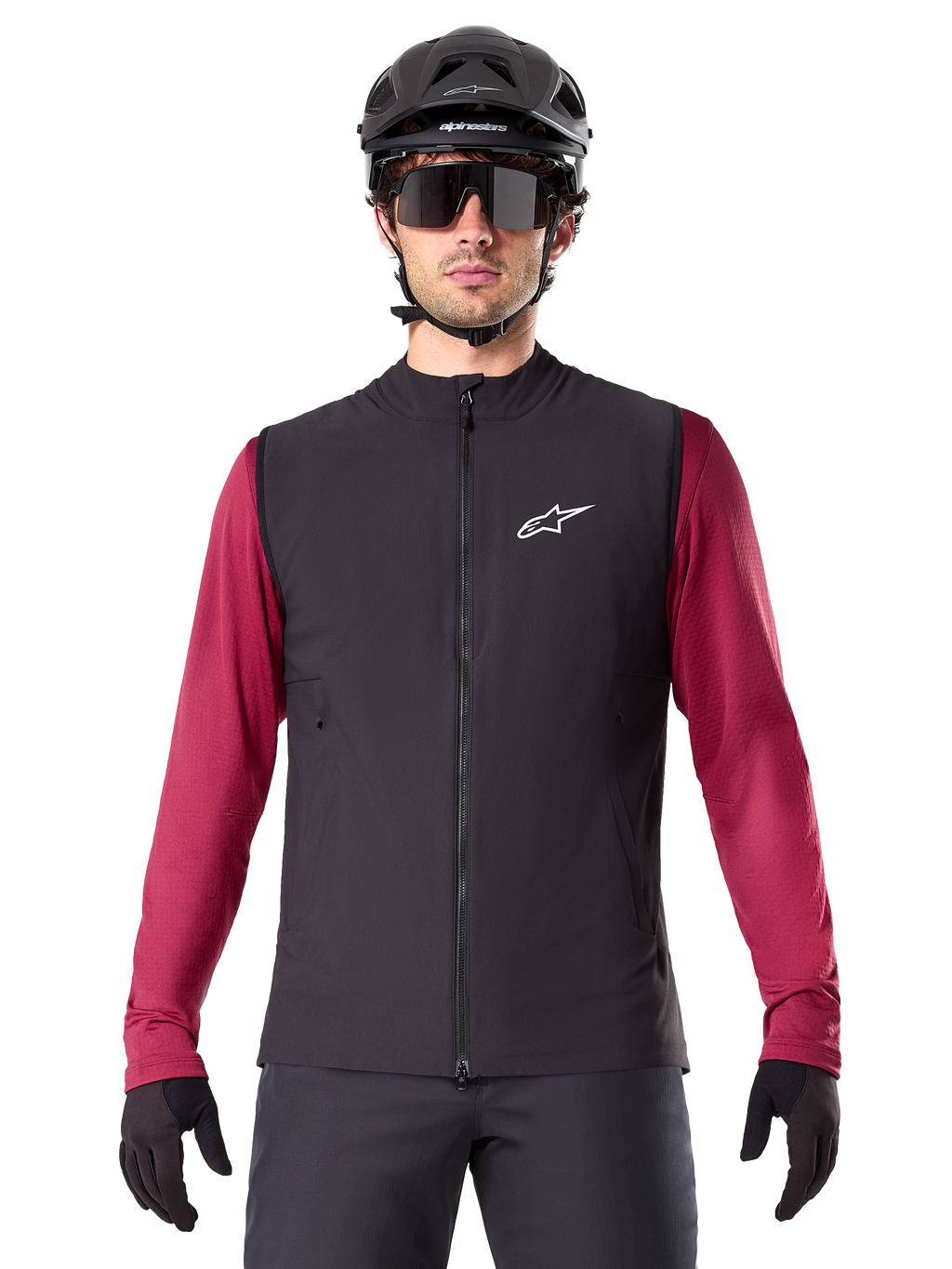 Alpinestars A-Dura Thermovest, zwart geïsoleerd fietsvest, gedragen door een model met een bordeauxrode jersey met lange mouwen, zwarte MTB-helm en zonnebril, voorzien van een wit logo op de borst, lichtgewicht thermische bescherming voor mountainbiken