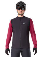 Alpinestars A-Dura Thermovest, zwart geïsoleerd fietsvest, gedragen door een model met een bordeauxrode jersey met lange mouwen, zwarte MTB-helm en zonnebril, voorzien van een wit logo op de borst, lichtgewicht thermische bescherming voor mountainbiken