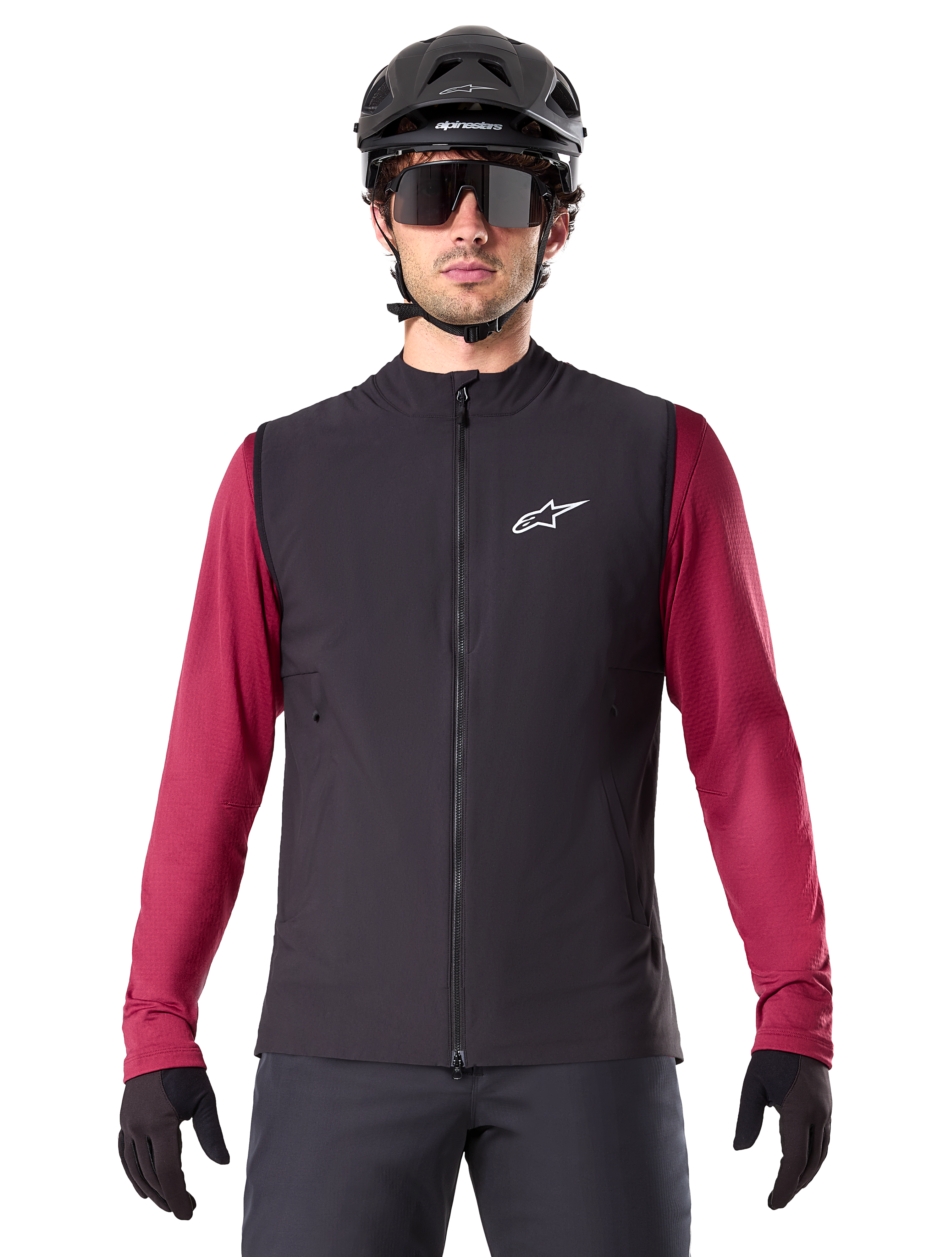 Alpinestars A-Dura Thermovest, zwart geïsoleerd fietsvest, gedragen door een model met een bordeauxrode jersey met lange mouwen, zwarte MTB-helm en zonnebril, voorzien van een wit logo op de borst, lichtgewicht thermische bescherming voor mountainbiken