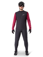 Alpinestars A-Dura Thermal Vest, zwart fietsvest gedragen over een bordeauxrode jersey met lange mouwen, gecombineerd met een donkergrijze MTB-broek, zwarte helm en zonnebril, technische mountainbikekleding voor ritten in koud weer