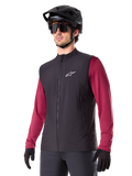 Alpinestars A-Dura Thermal Vest, zwart thermisch fietsvest, gedragen over een bordeauxrood shirt met lange mouwen, voorzien van een wit logo op de borst, gecombineerd met een zwarte MTB-helm en zonnebril, geïsoleerde prestatielaag voor mountainbiken