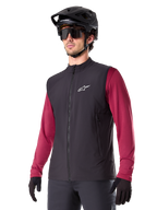 Alpinestars A-Dura Thermal Vest, zwart thermisch fietsvest, gedragen over een bordeauxrood shirt met lange mouwen, voorzien van een wit logo op de borst, gecombineerd met een zwarte MTB-helm en zonnebril, geïsoleerde prestatielaag voor mountainbiken