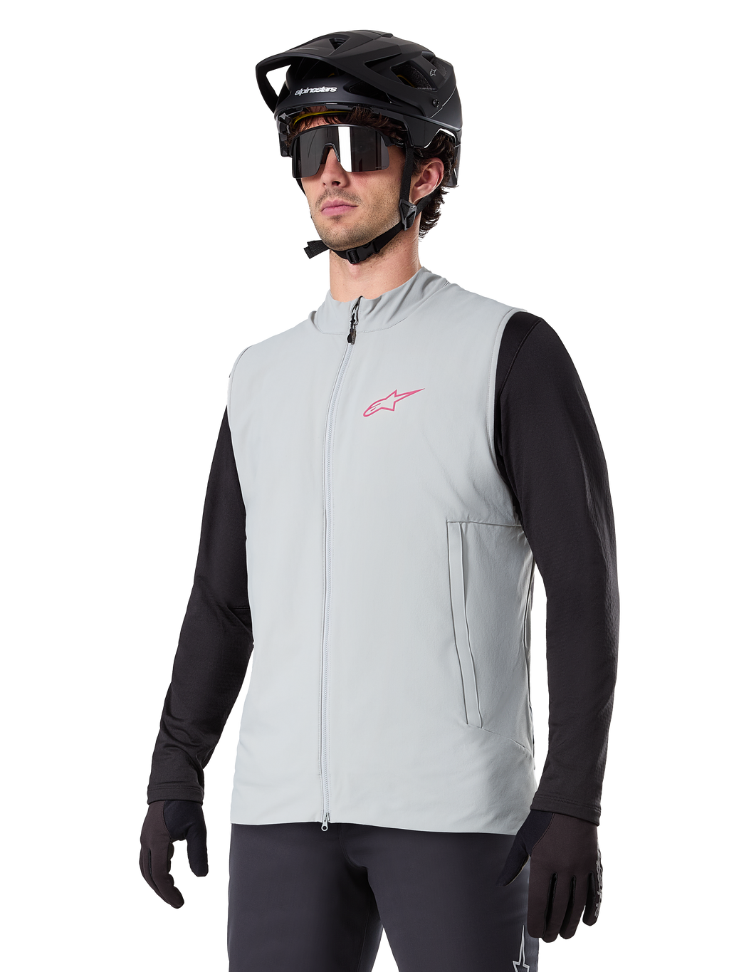 Alpinestars A-Dura Thermal Vest, Titanium grijs, geïsoleerd fietsvest, gedragen over een zwart shirt met lange mouwen met een zwarte mountainbikehelm en handschoenen, roze logo op de borst en verticale zijzakken met rits