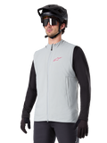 Alpinestars A-Dura Thermal Vest, Titanium grijs, geïsoleerd fietsvest, gedragen over een zwart shirt met lange mouwen met een zwarte mountainbikehelm en handschoenen, roze logo op de borst en verticale zijzakken met rits