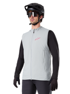 Alpinestars A-Dura Thermal Vest, Titanium grijs, geïsoleerd fietsvest, gedragen over een zwart shirt met lange mouwen met een zwarte mountainbikehelm en handschoenen, roze logo op de borst en verticale zijzakken met rits