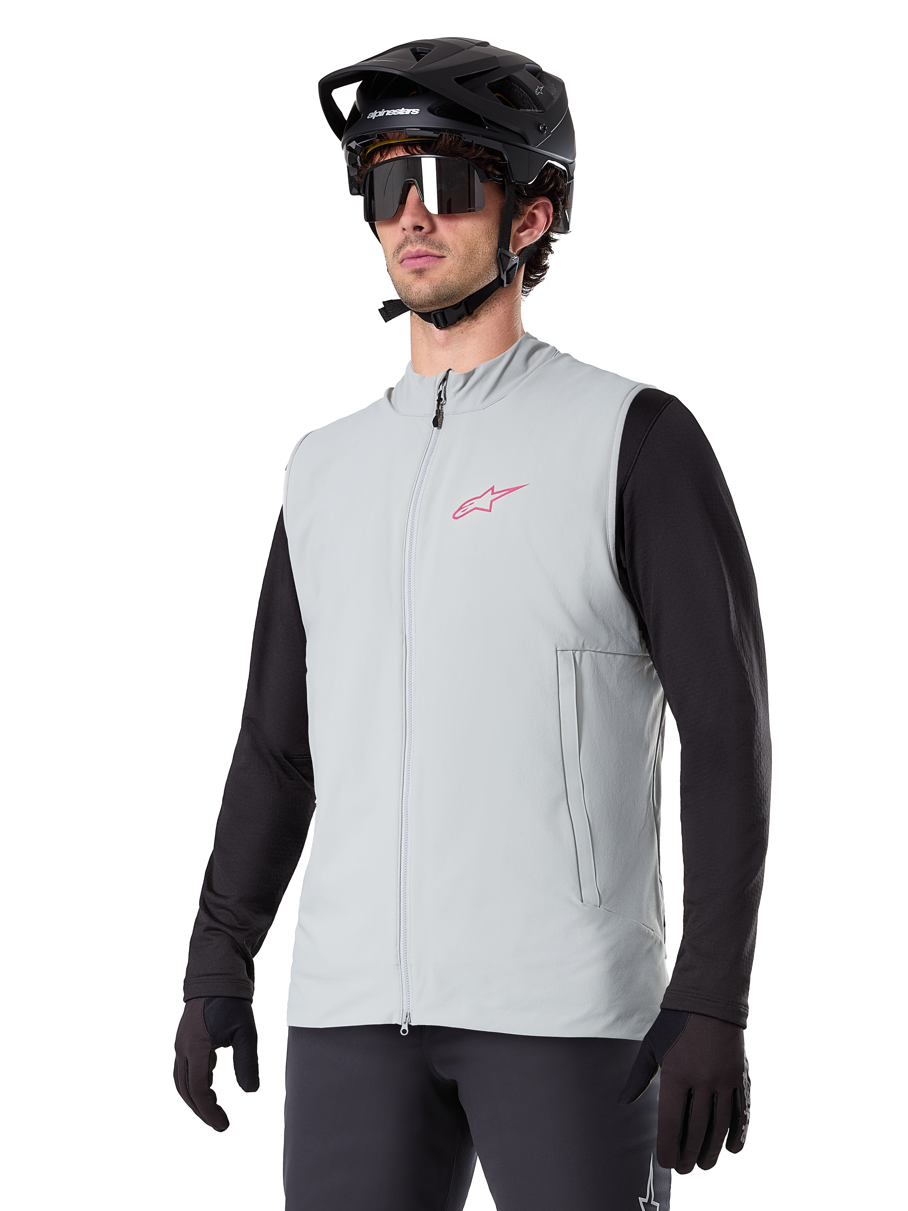 Alpinestars A-Dura Thermal Vest, Titanium grijs, geïsoleerd fietsvest, gedragen over een zwart shirt met lange mouwen met een zwarte mountainbikehelm en handschoenen, roze logo op de borst en verticale zijzakken met rits